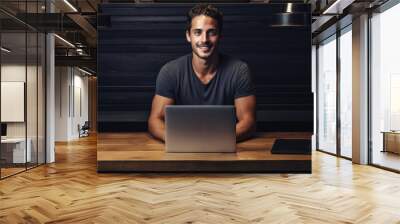 An attractive casual dressed man sitting at a desk with a laptop and smiling at the camera with a dark navy blue black wall behind him. Generative AI Wall mural