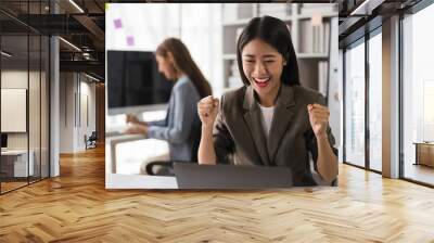 Businesswoman reading finance data on laptop and raising arm to celebrate with successful business Wall mural