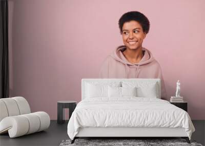 Happy attractive black girl using smartphone. Portrait with copy space. Cheerful smiling young African woman wearing casual hoodie with cell phone while standing over pink background Wall mural