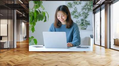 A young Japanese woman in her early thirties, wearing blue casual attire and smiling as she types on an open laptop at the white desk with a green plant behind it. She has short hair tied back  Wall mural