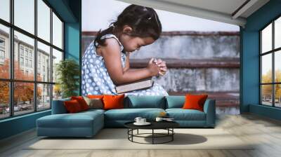 cute asian little girl closed her eyes and folded her hand in prayer on a holy bible for faith conce Wall mural