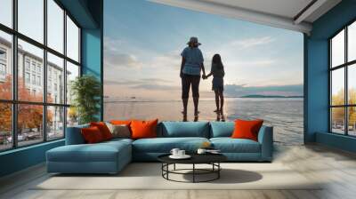 Silhouette of asian mother and daughter holding hand together standing on the beach looking at the beautiful sea and sky at the sunset time. Wall mural