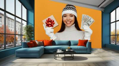 Christmas presents. Woman portrait. Accessories. Asian girl in a white polo neck, cap and gloves is holding a gift box and money and smiling, on an orange background Wall mural