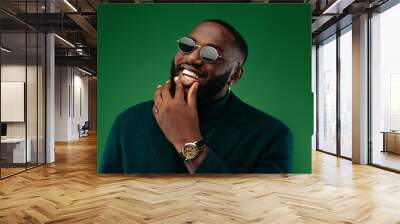 Man portrait. Style. Handsome Afro American guy in green jacket and sun glasses is looking at camera and smiling, on a green background Wall mural