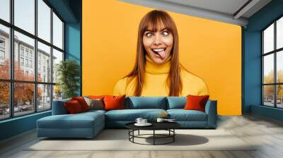 Woman portrait. Fun. Cheerful young woman is grimacing and showing her tongue at camera, on a yellow background Wall mural