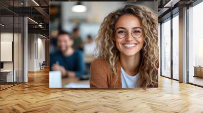 A cheerful woman with curly hair and glasses is smiling warmly, sitting in a vibrant, casual indoor setting with blurred people in the background. Wall mural