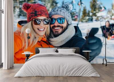 A couple dressed warmly in colorful winter attire enjoys a snowy day on the mountain, with ski lifts and other people skiing and sledding visible in the background. Wall mural