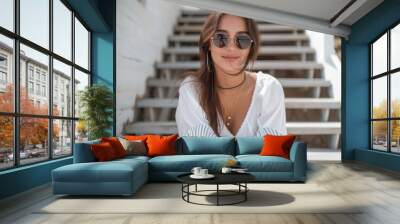 A fashionable woman confidently sits on the stairs, sporting a radiant smile and stylish sunglasses, embodying a perfect blend of chic clothing and functional eyewear Wall mural