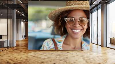 A fashionable woman with curly hair, wearing a stylish hat and sunglasses, smiles brightly while seated in a car, radiating warmth and a sense of adventure and confidence. Wall mural