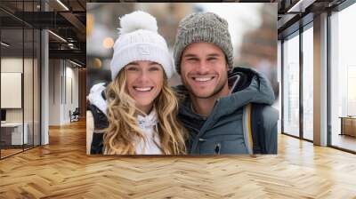 A joyful couple dressed in cozy winter clothing, embracing each other while smiling on a snowy city street, with an out-of-focus urban background featuring lights and trees. Wall mural