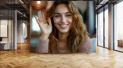 A joyful young woman is greeting with a hand wave and cheerful smile in a comfortable indoor setting Wall mural