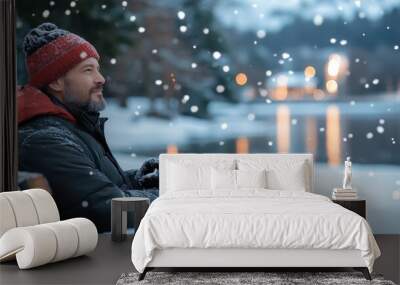 A man in cozy winter attire relaxes on a snowy park bench, surrounded by falling snowflakes and a tranquil lake view, embodying winter's peaceful essence. Wall mural