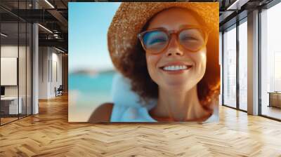 A serene woman in sunglasses and a straw hat enjoys the sun at the beach, wearing a bright smile, embodying a moment of pure relaxation and happiness. Wall mural