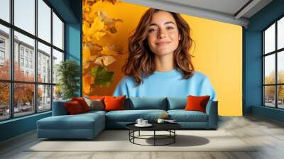 A smile lights up a woman's face as she poses with confidence in a blue sweater, set against a backdrop of rich, golden autumn foliage. Wall mural