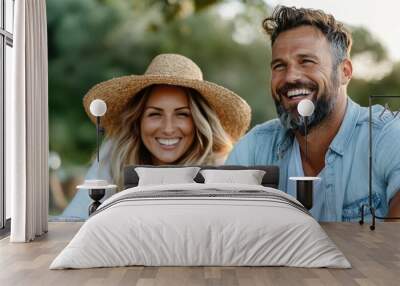 A smiling couple, the woman wearing a sun hat, sitting outdoors on a sunny day, enjoying a relaxed time together in a beautiful park setting with drinks. Wall mural