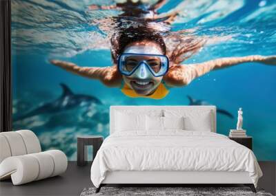 A woman diver, equipped in yellow swimwear, swims underwater alongside two dolphins in a vibrant, clear blue sea, showcasing a mesmerizing moment of nature and adventure. Wall mural