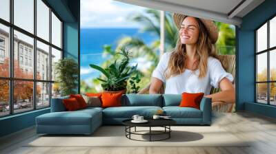 A woman in a straw hat smiles while sitting on a patio with tropical plants, overlooking the ocean, capturing a moment of serenity and natural beauty. Wall mural