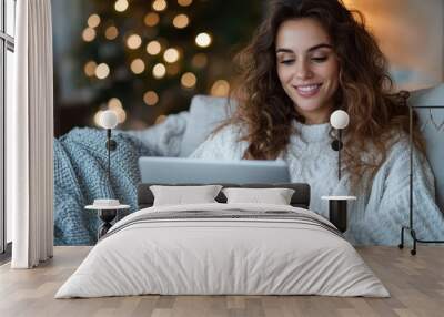 A woman in winter attire relaxes on a comfortable couch with a laptop, illuminated by warm ambient lights that create a festive and cozy home setting. Wall mural