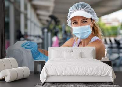 A woman is seen cleaning the railing in a gym while wearing protective gloves and a hair cover, highlighting the importance of hygiene and cleanliness in public spaces. Wall mural