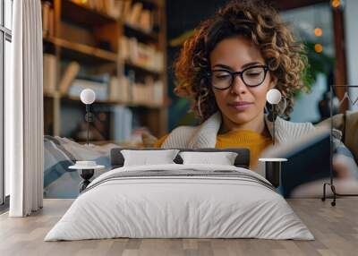 A woman with curly hair and glasses is intently reading a book while sitting in a comfortable, well-lit living room with a bookshelf in the background. Wall mural