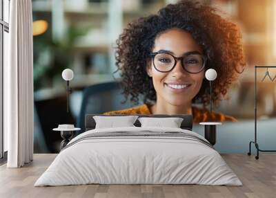 A woman with curly hair and glasses is seen joyfully smiling while sitting at her desk with sunlight streaming in, creating a motivating and positive work environment atmosphere. Wall mural