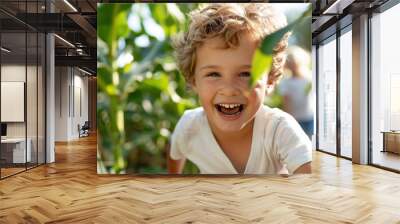 A young boy with curly hair laughs joyfully amidst lush green corn plants, capturing a heartwarming and spirited moment of fun and adventure in a natural setting. Wall mural