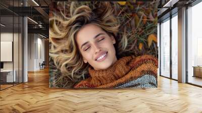 Blonde woman dressed in a warm crocheted sweater, lying on a grassy field with her eyes closed, surrounded by autumn leaves, capturing a serene and happy moment in nature. Wall mural