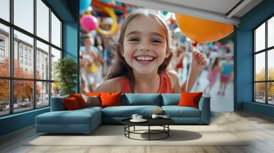 Enthusiastic young girl with long hair holds colorful balloons, grinning widely in a lively festival atmosphere with various people having fun in the background. Wall mural