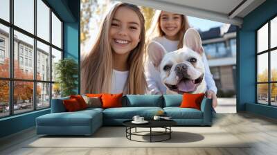 Two young sisters smile happily with their French Bulldog outside a suburban home, highlighting the joy of companionship and family moments in a sunny setting. Wall mural