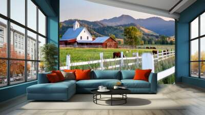 White barn and a few cows grazing in a lush green valley. Framed by a white fence and backed by rolling hills and a distant mountain range under a bright afternoon sky. Wall mural