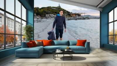 Portrait of young volunteer man looking at the camera while standing on the beach shore with barefoot holding a bag full of waste. Wall mural