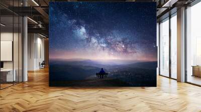 A man sitting on a bench staring at a starry sky with a Milky Way and a mountain landscape Wall mural