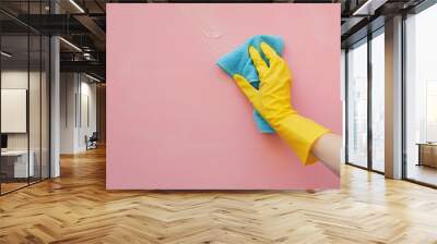 Employee hand in yellow rubber protective glove wiping pastel pink wall from dust with blue dry rag. General or regular cleanup. Commercial cleaning company. Copy space Wall mural