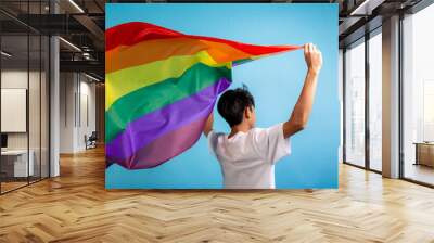 A man seen from behind holding colorful rainbow flag on blue color background. Male with a gay pride flag in studio, LGBT concept. Wall mural