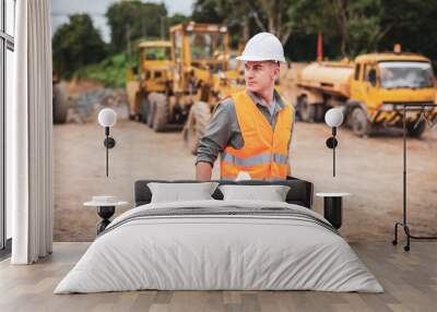 Caucasian handsome engineer is holding a laptop with roll of paper on road construction vehicle background. Engineer work concept Wall mural