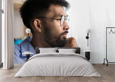 Thoughtful young bearded man sad in eyeglasses looking out window, serious employee thinking about project in the office. Wall mural