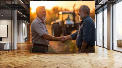 Two smiling men, one European and one African, shake hands warmly in a golden field at sunset, with a tractor in the background. Wall mural