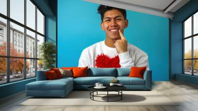 Young boy smiling and wearing a white sweat with a big red heart, pointing his chin with his finger Wall mural