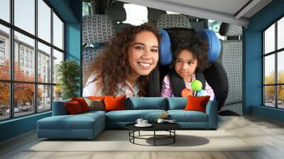 African-American woman and her little daughter with toy fastened in car safety seat Wall mural