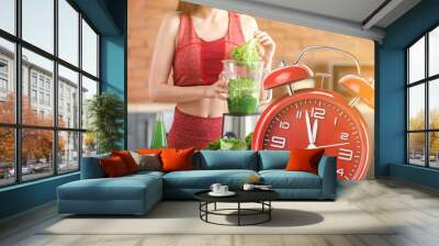 Alarm clock and young sporty woman preparing healthy vegetable juice in kitchen. Time to be healthy Wall mural