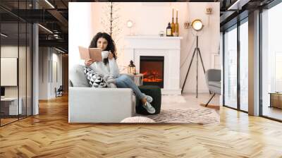 Beautiful woman drinking tea and reading book near fireplace at home Wall mural