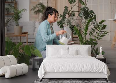 Beautiful young African-American woman watering plants while sitting on floor in living room Wall mural