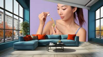 Beautiful young Asian woman applying cosmetic grape seed oil on lilac background Wall mural