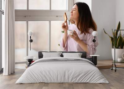 Beautiful young Asian woman drinking coffee and making tasty toasts in kitchen Wall mural