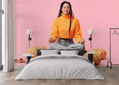 Beautiful young happy African-American woman with pumpkins and autumn leaves meditating on pink background Wall mural