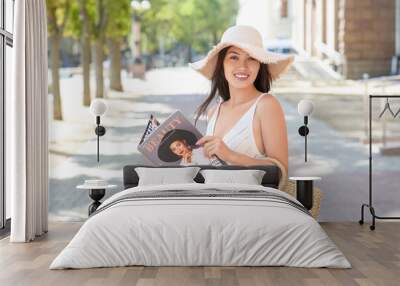 Beautiful young happy Asian woman in stylish wicker hat with bag reading magazine on city street Wall mural