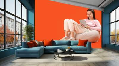 Beautiful young happy woman with tablet and cup of tea resting on beanbag chair against orange background Wall mural