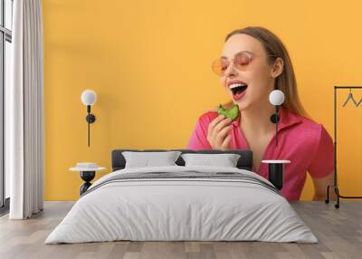 Beautiful young woman eating green sweet macaroon on yellow background Wall mural