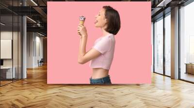 Beautiful young woman eating sweet ice-cream in waffle cone on pink background Wall mural