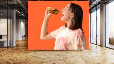 Beautiful young woman eating tasty sandwich on orange background Wall mural